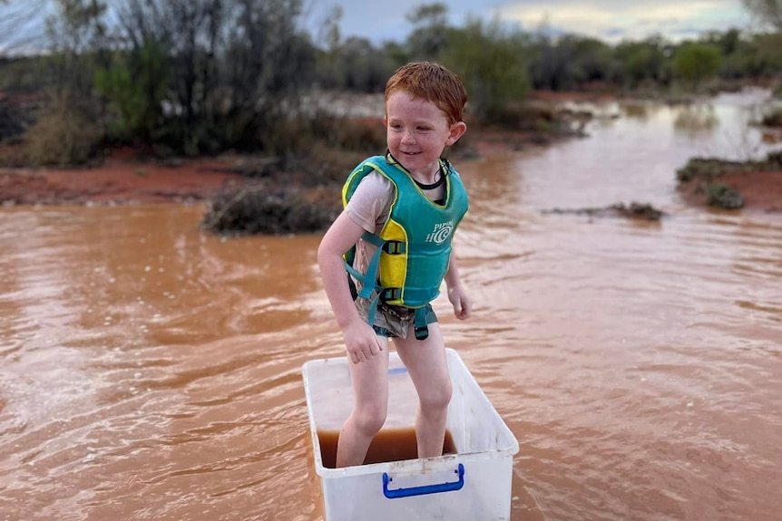 a young kid stands in a plastic tub floating in a big puddle