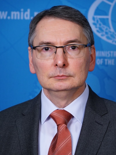 A clean shaven, middle aged man with short hair wears spectacles as he poses for a headshot in front of a blue background.