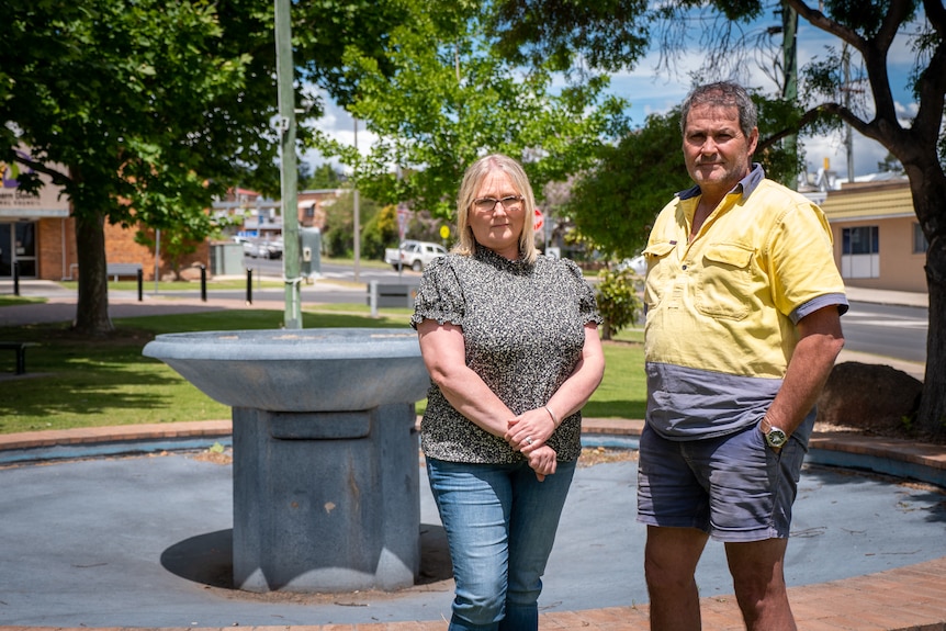 A woman wearing a black and white t-shire and a man wearing a yellow high-vis t-shirt stand before a water fountain in a park.