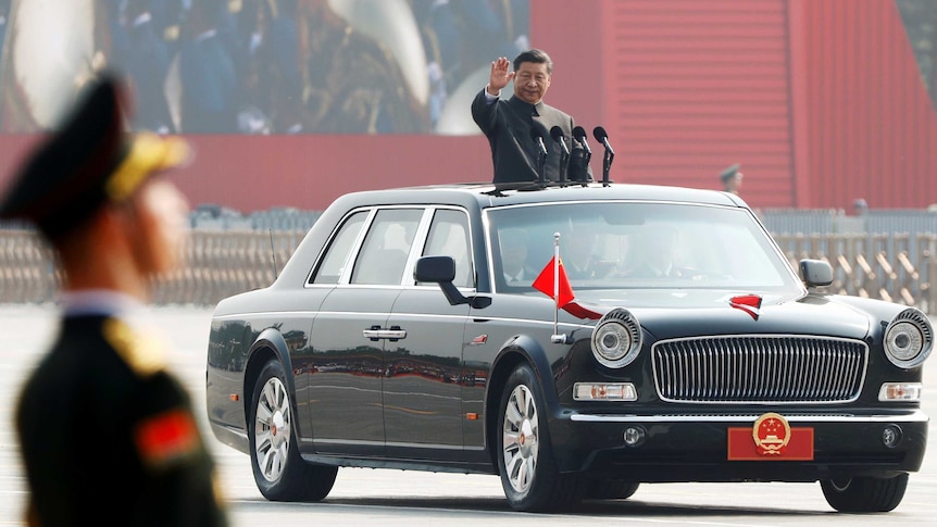 Xi Jinping waves from a black vehicle as it drives through a parade of some 15,000 troops.