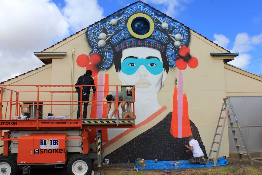 The mural in progress. One student and one teacher are working on a scissor lift while Fin DAC paints on the ground.