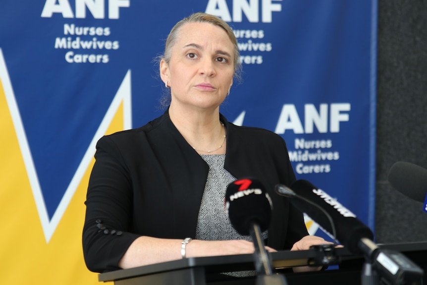 A woman stands at a lectern looking serious as she listens to a question. 