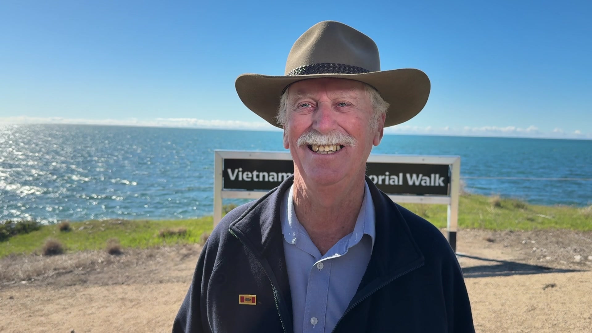 A Vietnam War veteran stands smiling in front of the Vietnam Memorial Walk sign.