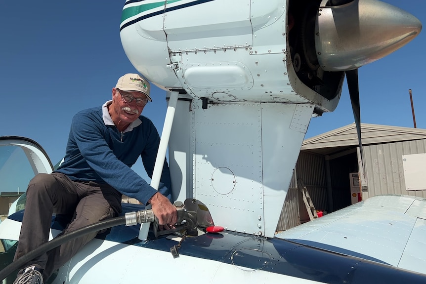 A man in a hat sits on the wing, refueling the plane, with a cabin in the background, a sunny day, the pilot is looking at the camera