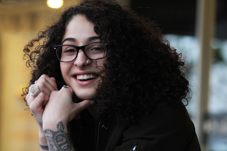 A young woman with dark curly hair looks straight at the camera, smiling.