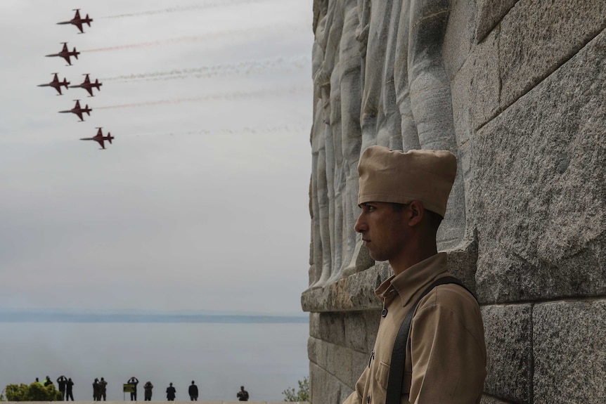 A man stands in profile in a brown uniform looking out as fighter jets fly in the sky.