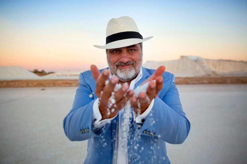 A man stands at a salt harvesting site, he holds handfuls of the salt and the flakes are falling to ground