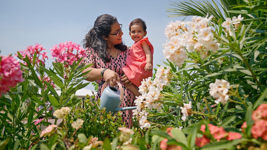 A woman with black hair wearing a pink dress smiles at her baby daughter in a garden
