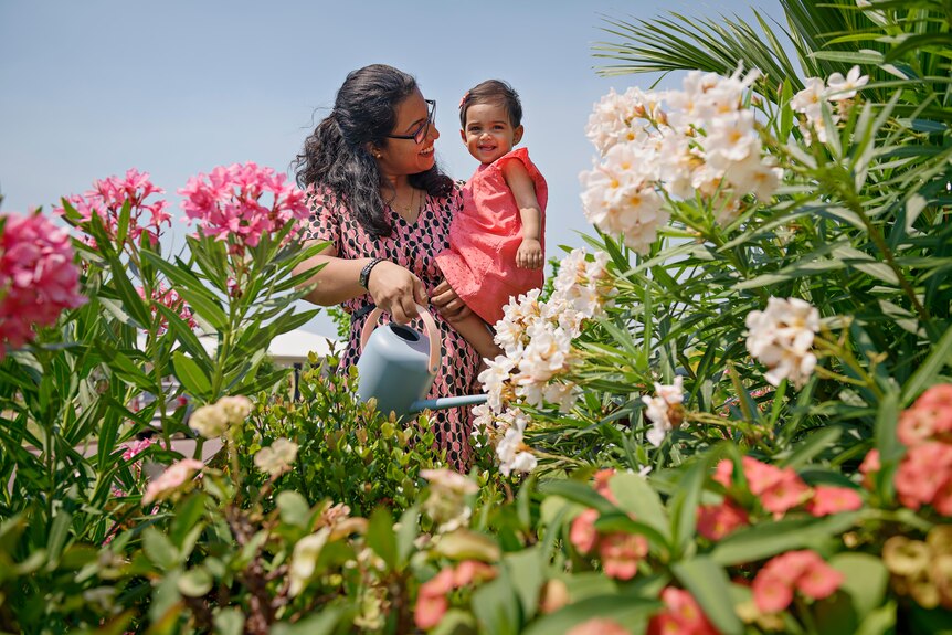 A woman with black hair wearing a pink dress smiles at her baby daughter in a garden