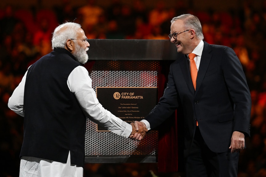 Two men shake hands in front of a plaque