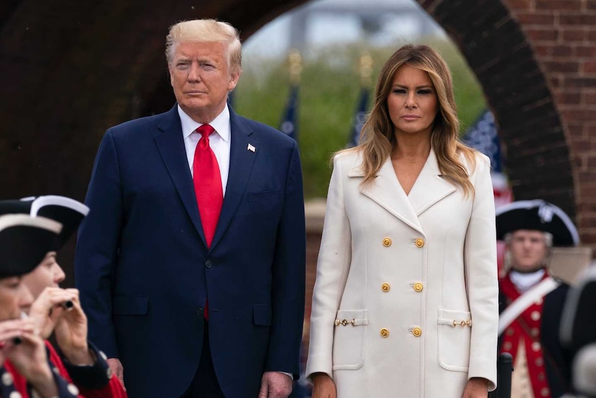 Blonde haired man wearing dark blue suit and red tie stands next to brunette woman in long white coat. Either side are soldiers