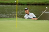 A golfer looks up at the ball as sand sprays from his shot from a bunker.