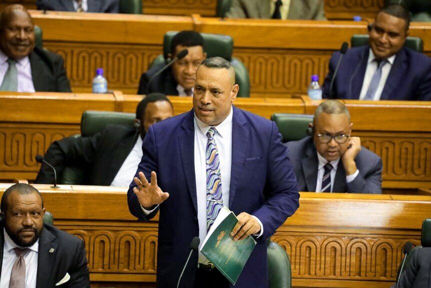 A man in blue suit and striped tie stands in a parliamentary chamber with wooden benches