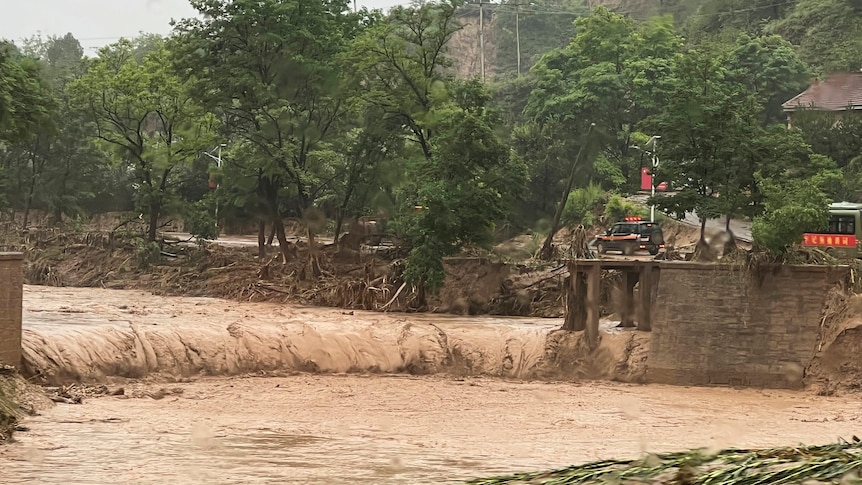 Car beside remnant of a bridge that was washed away by flash flooding, as muddy waters flow along the river.