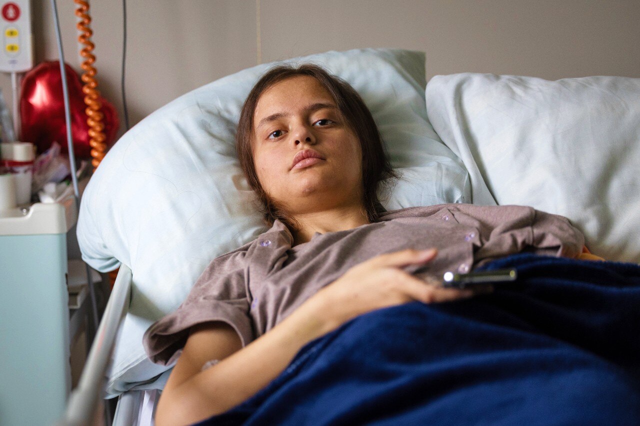 A woman lying in a bed, looking at the camera with a neutral expression on her face.