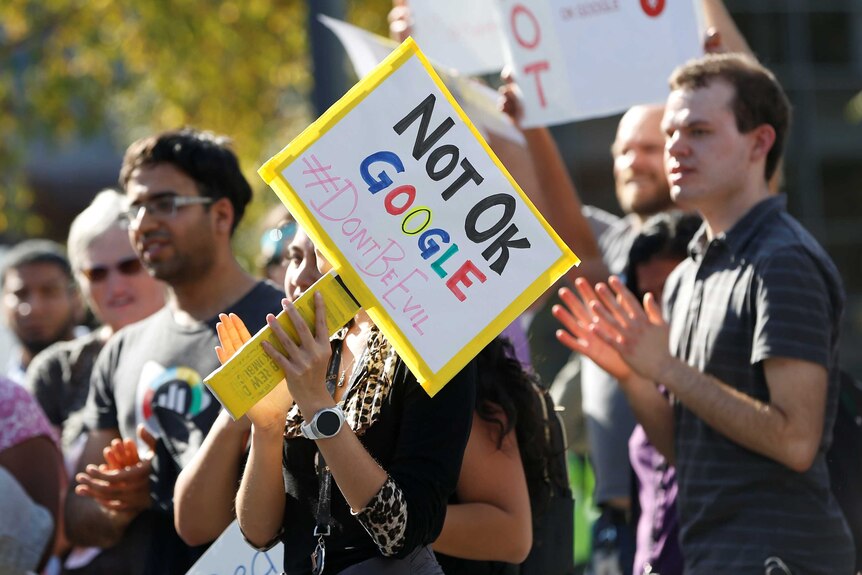 A sign saying Not OK Google #Dontbeevil is held up in front of a crowd of protesters