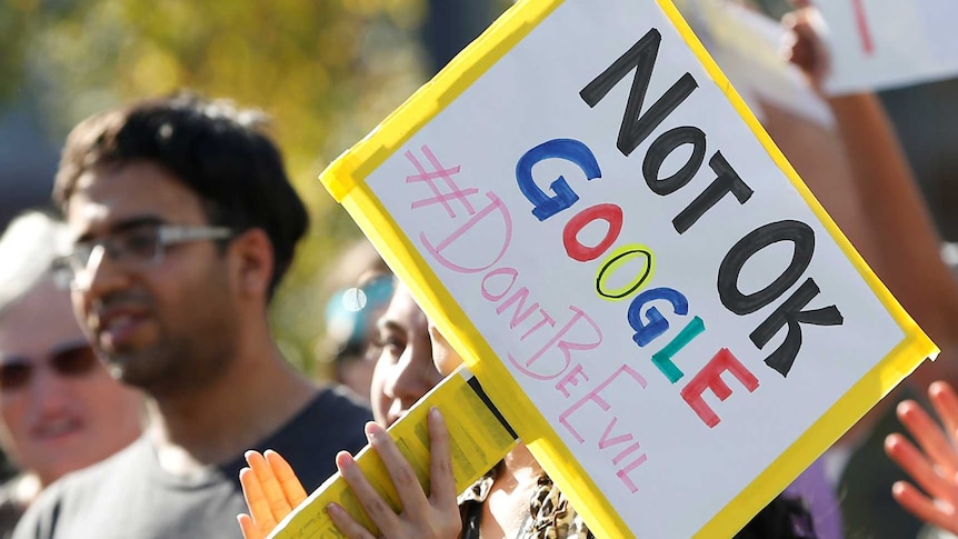 A sign saying Not OK Google #Dontbeevil is held up in front of a crowd of protesters