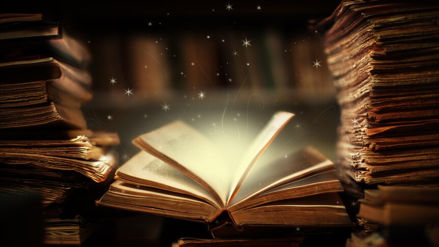 An open book sitting on a desk covered with papers and books. Stars and golden light is coming out of the book.