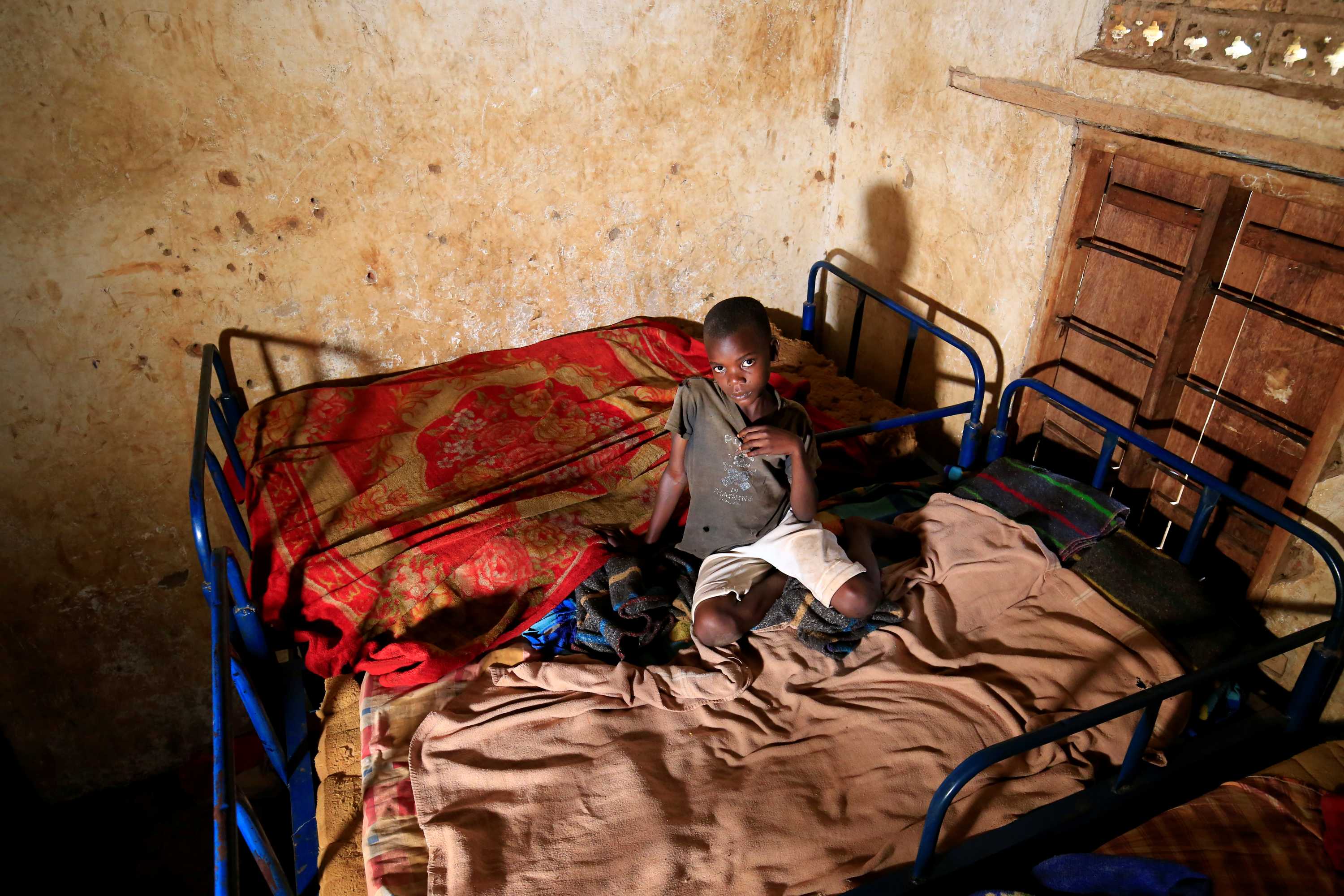 A child sits on two beds with thin mattresses pushed together, looking up at a camera.