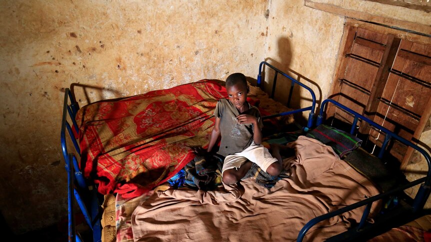 A child sits on two beds with thin mattresses pushed together, looking up at a camera.