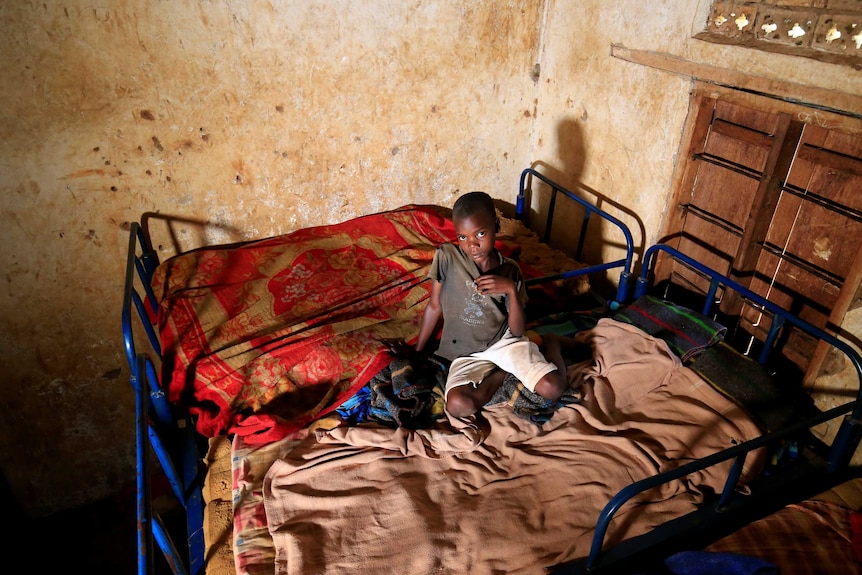 A child sits on two beds with thin mattresses pushed together, looking up at a camera.