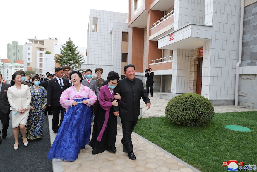 Женщина держит Ким Чен Ына за руку, в то время как группа людей следует за ним возле многоквартирных домов в дневное время.