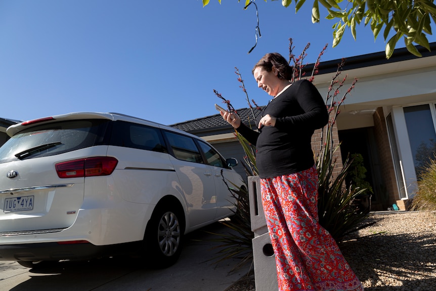 Tamika Cefai walks outside her house next to her drive way looking down at her phone on a sunny blue sky day.