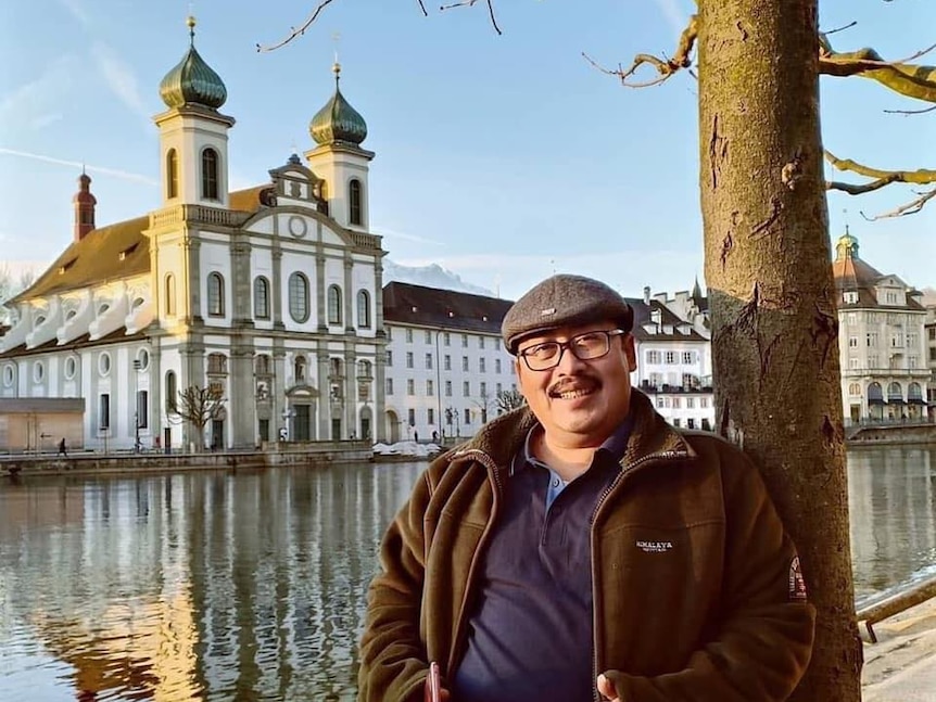 A man with a hat and glasses leans against a tree posing in front of a historic building.