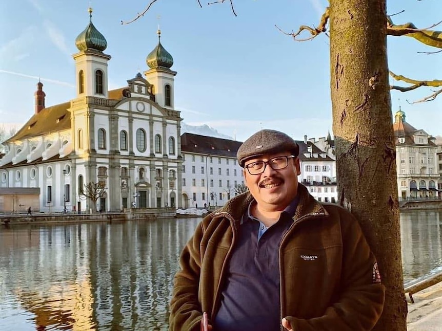 A man in a hat and glasses leans against a tree posing in front of a historic building.