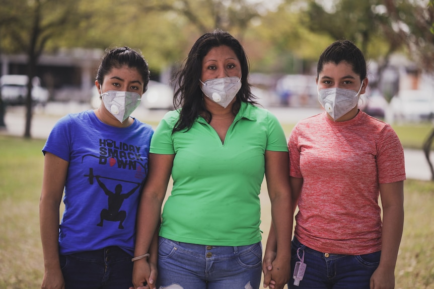 A woman with black hair and wearing a green shirt and mask stands next two two women with black hair wearing masks