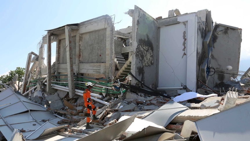 A man in an orange suit wearing a helmet inspecting rubble where a hotel used to stand.