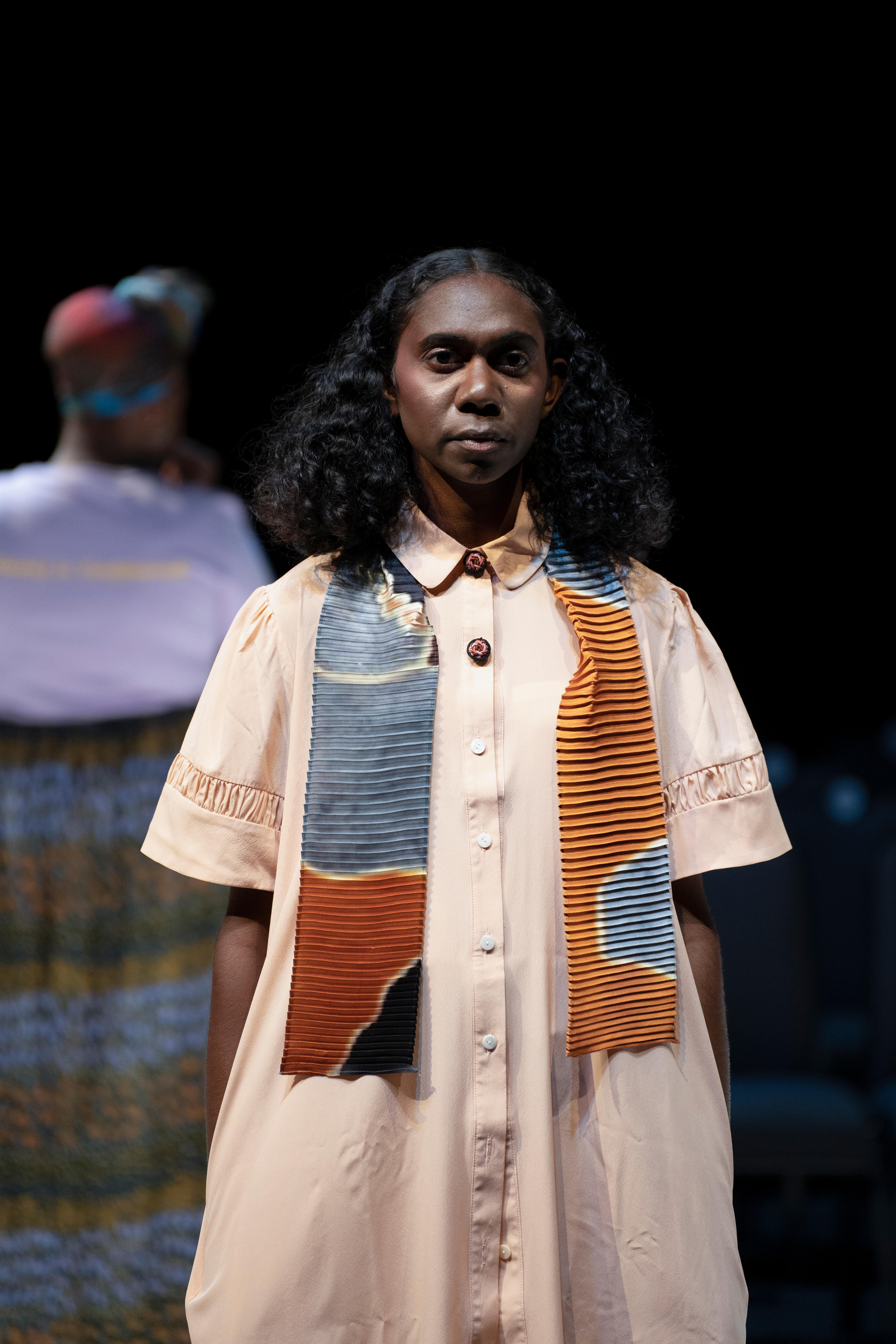 A woman wears a colorful scarf and looks into the camera against a black background. 