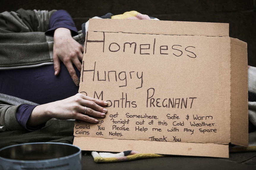 A pregnant woman lies on a footpath with a sign which says she is homeless, hungry, cold and asks for spare coins or notes.