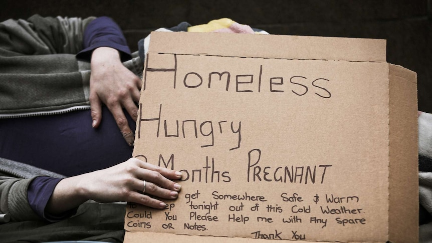 A pregnant woman lies on a footpath with a sign which says she is homeless, hungry, cold and asks for spare coins or notes.