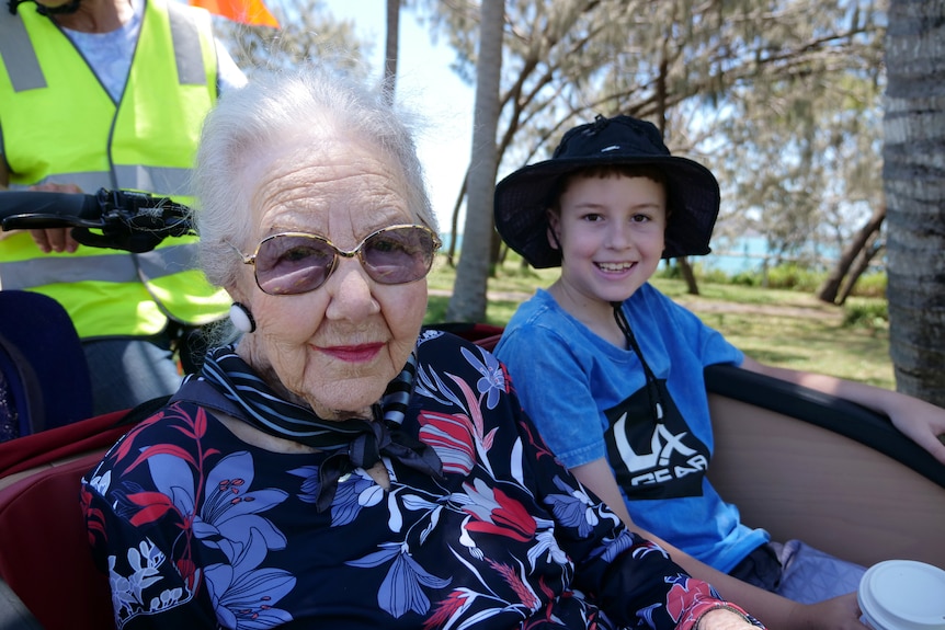 Thelma wearing glasses and lipstick sitting next to Max, wearing a blue shirt and a hat, smiling.