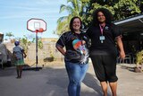 Two women stand in a courtyard while a young Indigenous boy plays basketball
