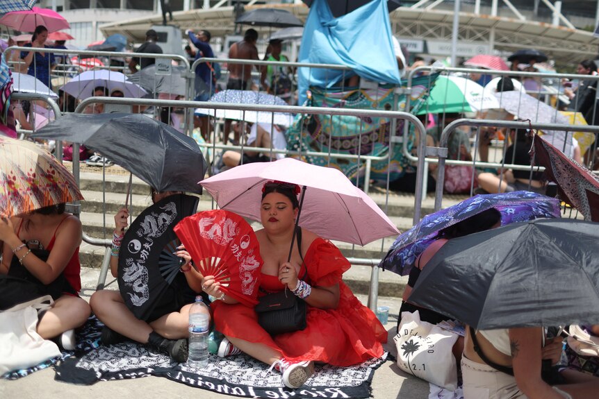 A girl in a red dress sits in a queue holding a fan under an umbrella 