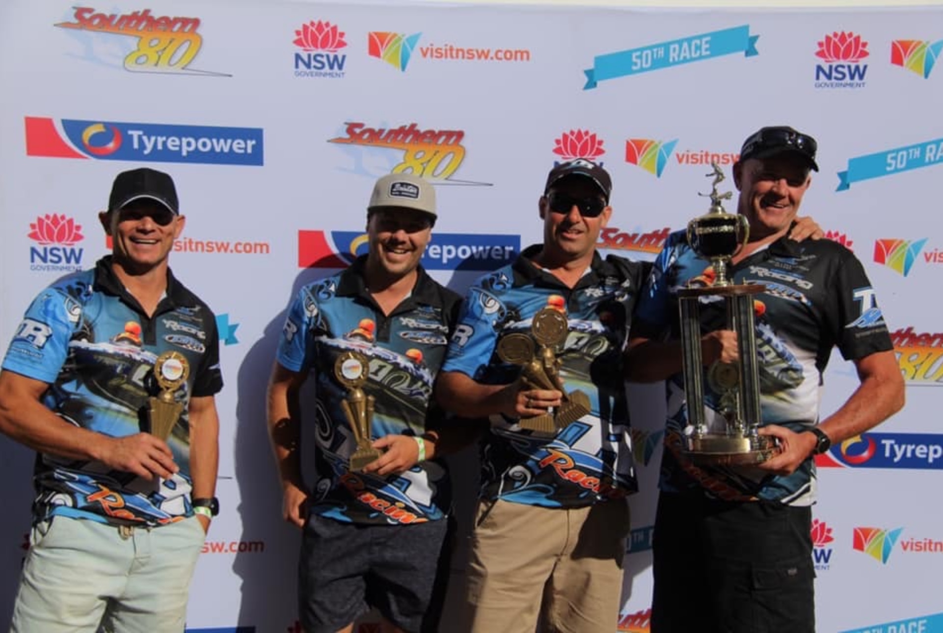 Cuatro hombres con camisetas del mismo equipo sostienen trofeos. 
