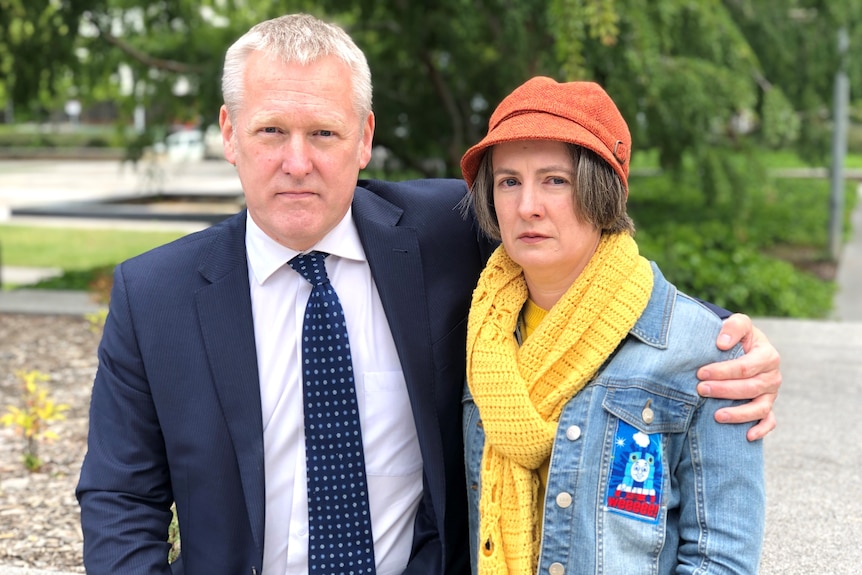 A man and a woman stand side by side with serious looks on their faces.