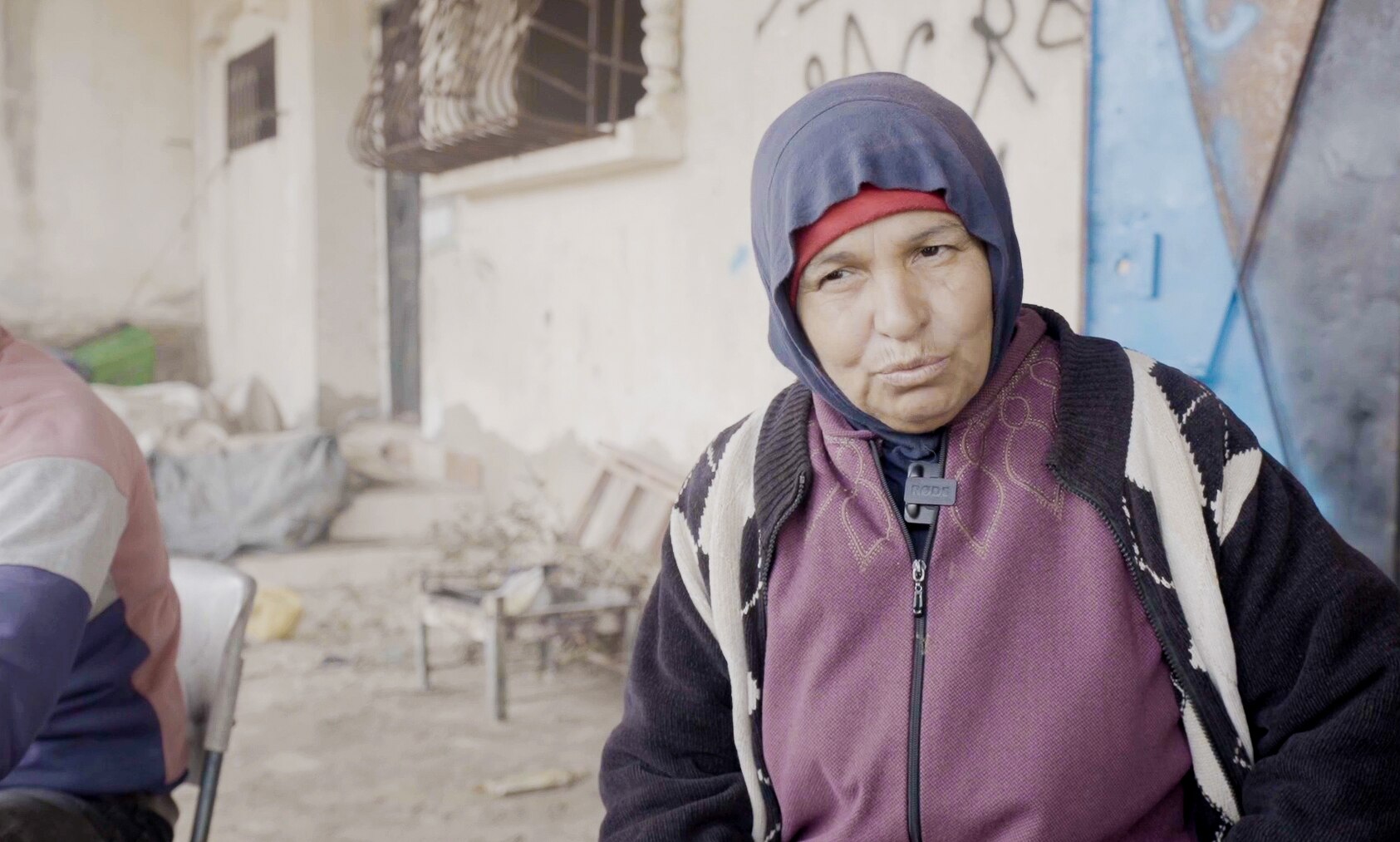 A close up of a woman wearing a red vest and head covering standing in front of a damaged building.