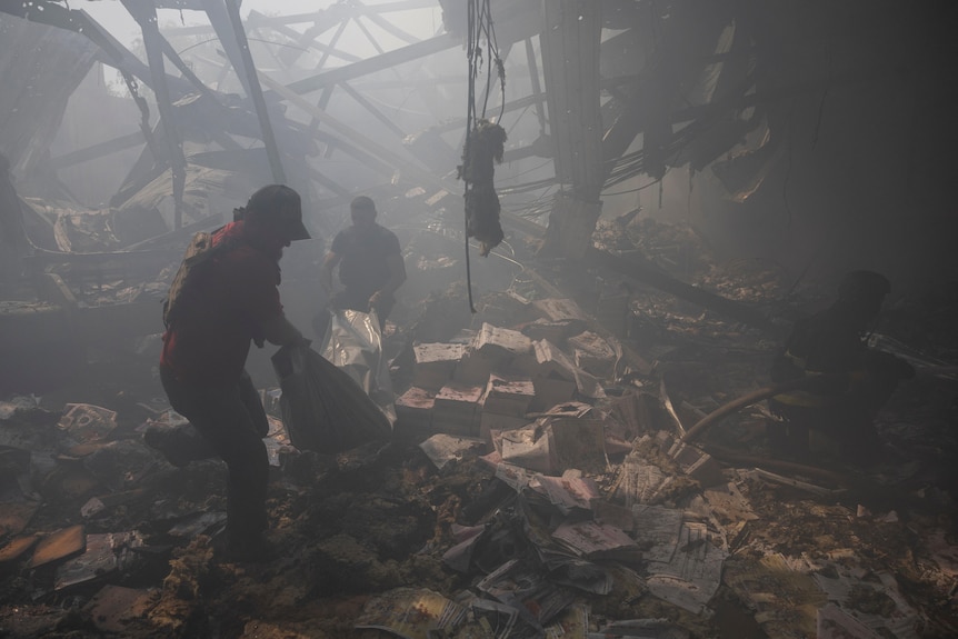 Two people lift a body bag out of the ruins of a building over stacks of charred paper