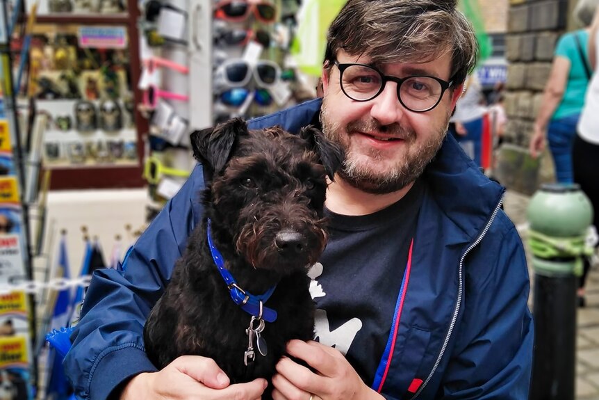 A close up photo of a man with a salt and pepper beard and wearing glasses holding a dog in a shop.