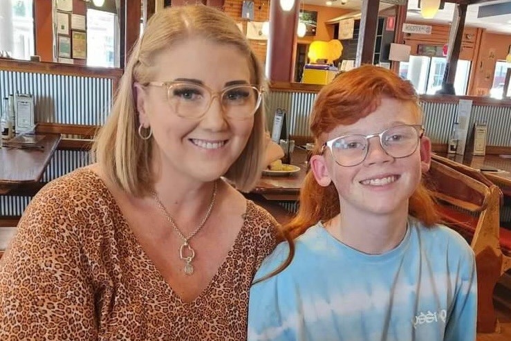 A smiling woman with blonde hair and glasses with a young boy sporting a long red mullet hairstyle.