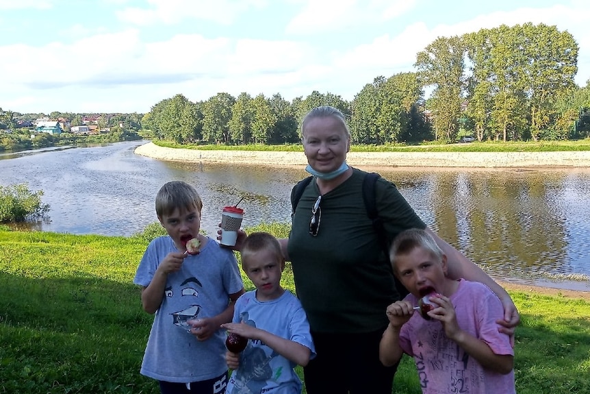 A smiling mother holds her arms around her three blond sons as they snack on toffee apples on a verdant river bank in sunlight.