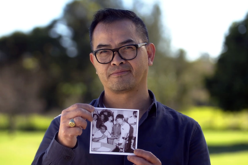 A vietnamese man with glasses holds a black and white photo showing him at age 14 aboared a boat
