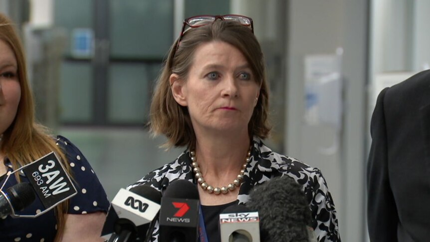 A woman with glasses sitting up on her head addresses a group of journalists in a hospital corridor.