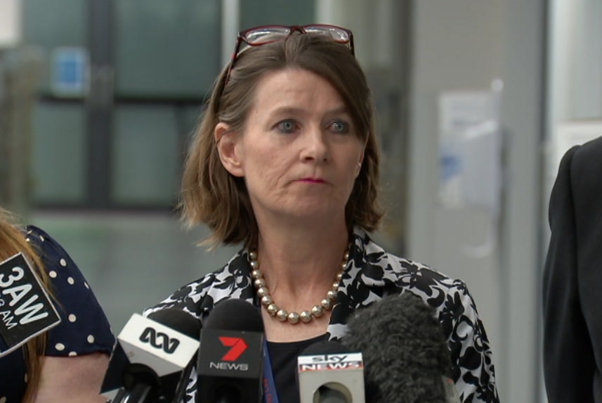 A woman with glasses sitting up on her head addresses a group of journalists in a hospital corridor.