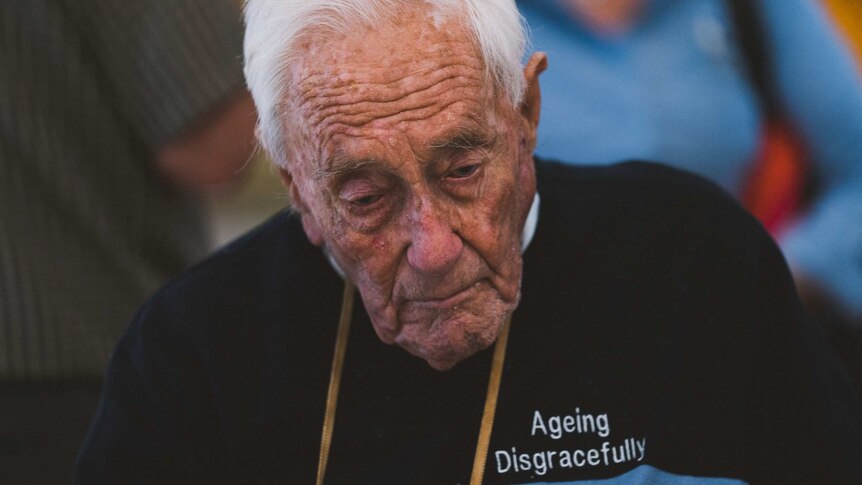 A close-up shot of an elderly man wearing a blue jumper with the words "ageing disgracefully" on the breast.