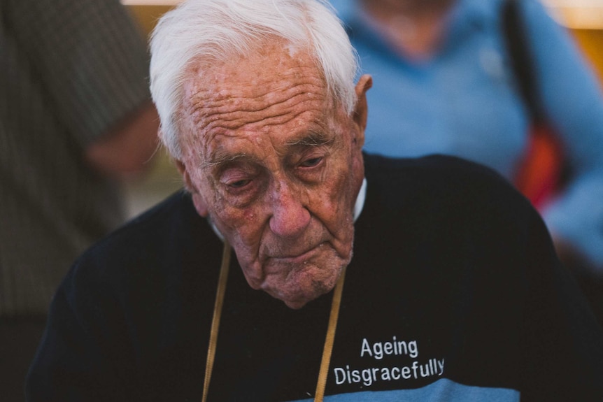 A close-up shot of an elderly man wearing a blue jumper with the words "ageing disgracefully" on the breast.