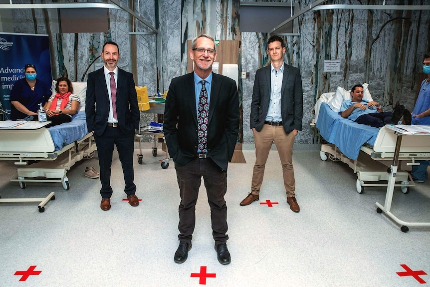 Three men standing amid a hospital ward with patients and nurses either side.